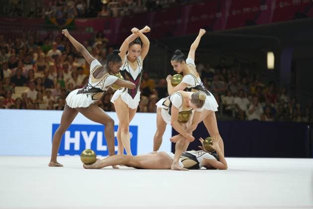 Eine Gruppe von fünf Turnerinnen führt eine synchronisierte Gruppenübung mit goldenen Bällen auf dem weißen Wettkampfteppich der Festhalle Frankfurt durch. Mehrere Athletinnen strecken die Fäuste nach oben, während eine Turnerin am Boden liegt und eine weitere sich tief bückt. Gruppenübung mit goldenen Bällen bei der Rhythmischen Sportgymnastik WM in Frankfurt *** A group of five gymnasts performs a synchronized group routine with golden balls on the white competition mat at the Festhalle in Frankfurt. Several athletes raise their fists in the air, while one gymnast lies on the floor and another bends down deeply. Group routine with golden balls at the Rhythmic Gymnastics World Championships in Frankfurt Copyright: xspeedshotx