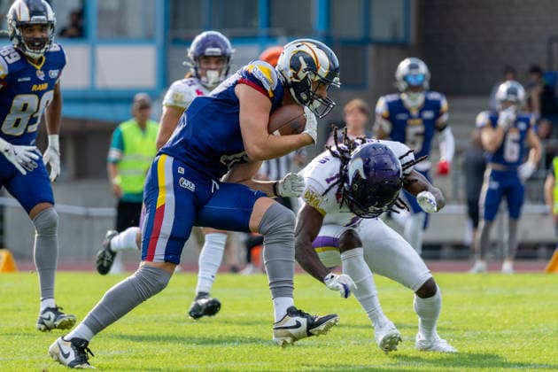 An Alpine Rams player advances with the football while driving forward through contact on the field. Nearby, a Vienna Vikings defender reaches out during the play while other players surround the action. Spectators and stadium seating fill the background.. Vienna Vikings at Alpine Rams, American Football, American Football League Europe, Week 13, 2026, 8.8.2026, Copyright: xPFA RicoxDoninellix
