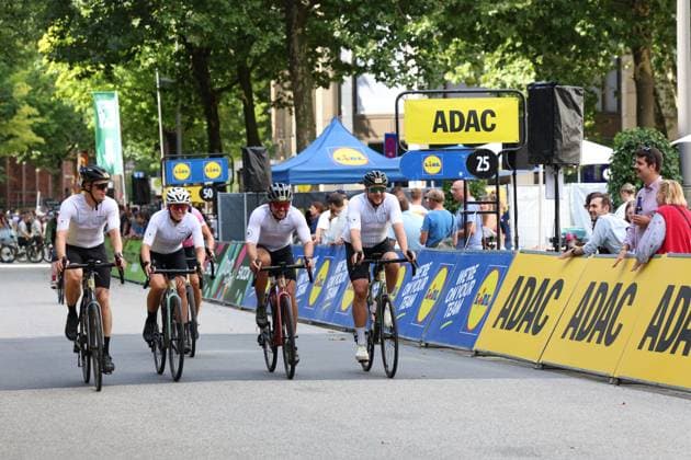 Vier Radfahrer in weißen Trikots fahren gemeinsam über die Ziellinie am Rathausmarkt Hamburg, flankiert von ADAC- und Lidl-Werbebanden. Im Hintergrund verfolgen zahlreiche Zuschauer das Rennen. Radfahrer passieren die Zieldurchfahrt bei den Cyclassics Hamburg *** Four cyclists in white jerseys cross the finish line together at Hamburg’s Rathausmarkt, flanked by ADAC and Lidl advertising banners. In the background, numerous spectators watch the race. Cyclists pass the finish line at the Cyclassics Hamburg. Copyright: xspeedshotx xSarahxWolfx