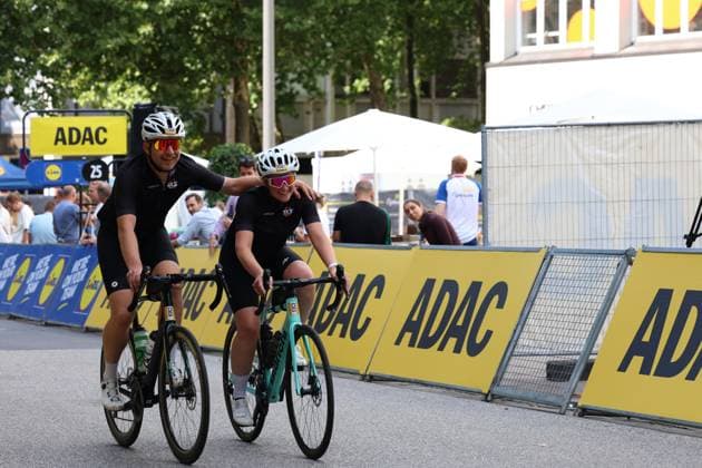 Zwei Radfahrer in schwarzen SKS-Trikots fahren nebeneinander am Rathausmarkt Hamburg, einer legt den Arm um die andere Person – beide lächeln in die Kamera. Im Hintergrund sind ADAC-Werbebanden und Zuschauer zu sehen. Zwei Radfahrer in schwarzen Trikots beim Cyclassics Hamburg am Rathausmarkt *** Two cyclists in black SKS jerseys are riding side by side at Hamburg’s Rathausmarkt; one has an arm around the other—both are smiling at the camera. ADAC advertising banners and spectators can be seen in the background. Two cyclists in black jerseys at the Cyclassics Hamburg race at Rathausmarkt Copyright: xspeedshotx xSarahxWolfx