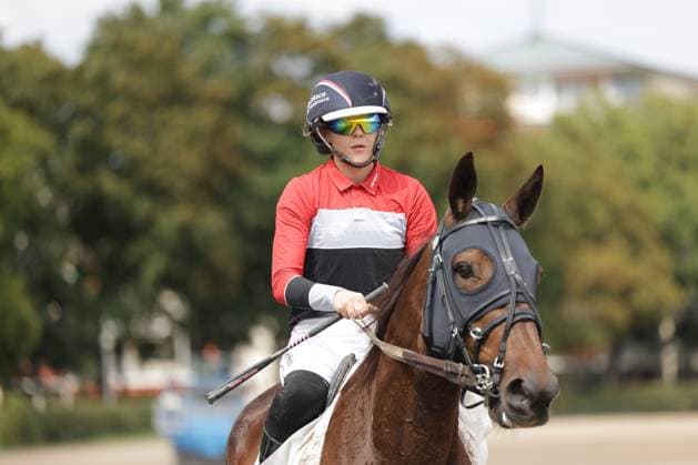 Eine Jockeyrin in rotem Dress mit Schutzhelm und Sportbrille reitet auf einem braunen Vollblüter über die Sandbahn der Rennbahn Berlin Mariendorf. Pferd und Reiterin sind scharf freigestellt vor einem grünen, unscharfen Baumhintergrund. Jockeyrin auf braunem Pferd bei der Rennbahn Berlin Mariendorf *** A female jockey in a red outfit, wearing a helmet and sports glasses, rides a bay Thoroughbred across the sand track at the Berlin Mariendorf Racetrack. The horse and jockey are sharply focused against a green, blurred background of trees. Female jockey on a brown horse at the Berlin Mariendorf Racetrack Copyright: xspeedshotx