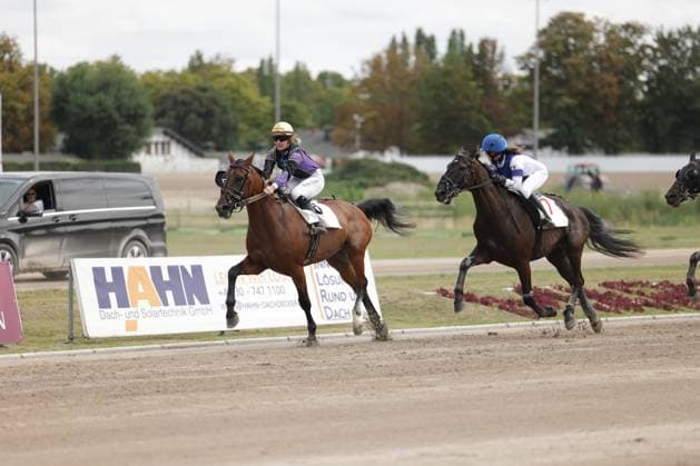Zwei Jockeys liefern sich ein enges Rennen auf der Sandbahn in Berlin Mariendorf, das führende braune Pferd mit lila-weißem Sattel knapp vor dem dunklen Pferd mit blau-weißem Sattel. Im Hintergrund sind Werbebanden und Bäume zu sehen. Jockeys im Kopf-an-Kopf-Rennen auf der Rennbahn Berlin Mariendorf *** Two jockeys are locked in a close race on the dirt track in Berlin Mariendorf, with the leading bay horse, bearing a purple-and-white saddle, just ahead of the dark horse with a blue-and-white saddle. In the background, advertising banners and trees are visible. Jockeys in a neck-and-neck race at the Berlin Mariendorf racetrack Copyright: xspeedshotx