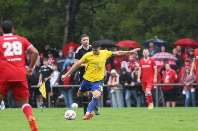 Semir Telalovi? ( 17; Rot-Weiss Essen) holt zum Schuss aus, während ein Spieler von SV Sonsbeck im Zweikampf hinter ihm ist. Im Hintergrund verfolgen zahlreiche Zuschauer mit roten Regenschirmen das Spiel im Willi-Lemkens Sportpark in Sonsbeck. Semir Telalovi? ( 17; Rot-Weiss Essen) im Schussmoment gegen SV Sonsbeck *** Semir Telalovi (17) of Rot Weiss Essen takes a shot while an SV Sonsbeck player challenges him from behind. In the background, numerous spectators with red umbrellas watch the game at Willi Lemkens Sportpark in Sonsbeck. Semir Telalovi (17) of Rot Weiss Essen takes a shot against SV Sonsbeck. Copyright: xspeedshotx xTimoxBluhmki-Schmidtx