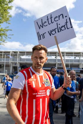 GER, Jena, Fussball, DFB-Pokal, 1. Hauptrunde, FC Carl-Zeiss Jena vs. SV Darmstadt 98 im Bild v.l.: Thomas Melchior , auf Social Media als Sportwettensheriff unterwegs, macht im Trikot des FC Rot-Weiss Erfurt und Spruchschild vor dem Stadion des FC Carl-Zeiss Jena auf Suchtpotenzial von Sportwetten aufmerksam , spricht mit Fans von Jena, geht in den Austausch 23.08.2026, Ernst Abbe Sportfeld Jena DFB REGULATIONS PROHIBIT ANY USE OF PHOTOGRAPHS AS IMAGE SEQUENCES AND OR QUASI-VIDEO, *** GER, Jena, Soccer, DFB Cup, First Round, FC Carl Zeiss Jena vs. SV Darmstadt 98: Pictured from left, Thomas Melchior, known on social media as the Sports Betting Sheriff, wearing an FC Rot Weiss Erfurt jersey and holding a sign in front of the FC Carl Zeiss Jena stadium to raise awareness about the addictive nature of sports betting, talking with Jena fans, engaging in conversation on August 23, 2026, Ernst Abbe Sportfeld, Jena. DFB REGULATIONS PROHIBIT ANY USE OF PHOTOGRAPHS AS IMAGE SEQUENCES AND OR QUASI-VIDEO, Copyright: HMBxMedia PaulxFritz