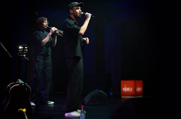 Zwischen Gewinner und Deja Vu Clueso in Stralsund: Ein Musiker erzählt von der Zeit und von sich selbst Es gibt Musiker, die betreten eine Bühne und verwandeln sie in einen Ort der großen Geste. Und es gibt Clueso. Bei ihm scheint das Gegenteil zu geschehen: Je größer die Karriere geworden ist, desto wichtiger wurde die Nähe. Heute Abend, im Theater Vorpommern in Stralsund, dürfte deshalb weniger der Popstar als vielmehr der Erzähler Clueso im Mittelpunkt stehen ein Musiker, der zwischen Liedern gern die Tür zu jener Welt öffnet, aus der diese Lieder stammen. stars@ndr2 songs & stories heißt das Format, das den gebürtigen Erfurter an diesem Montag ins Große Haus führt. Es ist ein bewusst intimer Gegenentwurf zur großen Konzertmaschine: Songs, Geschichten, Erinnerungen. Kein Stadion, kein spektakuläres Bühnenbild, sondern ein Künstler, der sein musikalisches Leben gewissermaßen noch einmal aufschlägt.Und dieses Leben reicht inzwischen weit zurück. 25 Jahre sind vergangen, seit Clueso mit seinem Debütalbum Text und Ton auf sich aufmerksam machte. Ein Vierteljahrhundert Popgeschichte und eine bemerkenswerte Entwicklung: vom jungen Rapper aus Erfurt zum Singer-Songwriter, dessen Lieder längst zum kollektiven Gedächtnis des deutschsprachigen Pop gehören.Dabei war Cluesos Weg nie der einer geradlinigen Karriereplanung. Seine musikalischen Wurzeln liegen im Hip-Hop und Rap. Aus dieser Herkunft hat er sich jedoch nicht verabschiedet, sondern sie in eine immer größere musikalische Offenheit überführt. Vielleicht erklärt das, warum seine Songs so selbstverständlich zwischen Sprechgesang, Pop, Soul und klassischem Songwriting wechseln können.Clueso ist ein Grenzgänger geblieben allerdings einer ohne demonstrative Pose. Seine großen Lieder funktionieren gerade deshalb, weil sie nicht so tun, als hätten sie die Welt bereits verstanden. Keinen Zentimeter, Gewinner, Chicago, später