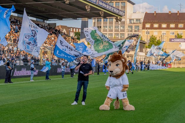 Sebastian Schaech (Stadionsprecher, TSV 1860 Muenchen) und Maskottchen Sechzgerl (TSV 1860 Muenchen) vor der Fankurve, im Hintergrund die Fahnenschwenker vor der Stehhalle GER, TSV 1860 Muenchen vs. TSV Schwaben Augsburg, Fussball, Regionalliga Bayern, 8. Spieltag, Saison 2026 2027, 11.09.2026. (DFL DFB REGULATIONS PROHIBIT ANY USE OF PHOTOGRAPHS as IMAGE SEQUENCES and or QUASI-VIDEO). Foto: HMB Media Heike Feiner Copyright: HMBxMedia HeikexFeiner