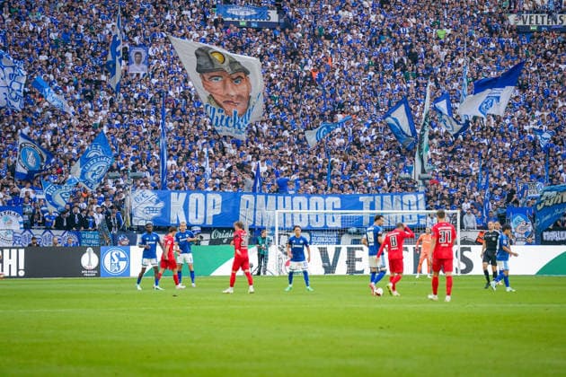 Deutschland: Fussball Bundesliga 2026 - 2027, 4. Spieltag - FC Schalke 04 vs SV Elversberg - Veltins-Arena (Gelsenkirchen): Fans des FC Schalke 04 in der dicht besetzten Nordkurve zeigen zahlreiche blau-weiße Fahnen und ein großes Transparent mit der Aufschrift KOP OMHOOG TIMO! (Kopf hoch, Timo!) während des Bundesliga-Spiels gegen den SV Elversberg. Der konkrete Hintergrund der Botschaft lässt sich aktuell nicht belastbar öffentlich verifizieren. Nordkurve, Fans, Ultras, Fankultur, Transparent, Spruchband, Solidarität, Fahnen, Hintertorperspektive, Totale. Copyright: xJOERANxSTEINSIEKx