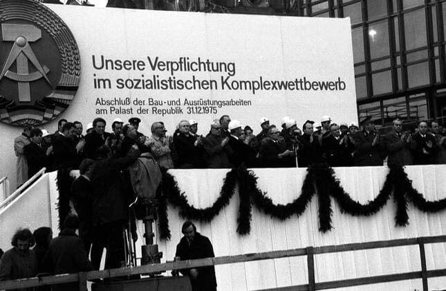 Unsere Verpflichtung im sozialistischen Komplexwettbewerb - Schriftzug auf einem Transparent hinter der mit klatschenden Genossen besetzten Tribüne während des Richtfestes am Palast der Republik in Ost Berlin