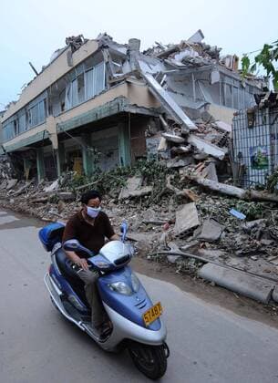 Einheimischer mit Mundschutz auf einem Motorroller vor einem zerstörten Haus in Qingchuan - 