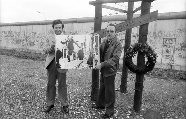Ikonische Bilder der Zeitgeschichte Conrad Schumann (li.) mit Peter Leibing 03 86 ru Der Hamburger Fotograf Peter Leibing (re.) schenkt im März 1986 dem Ex-DDR-Volkspolizisten Conrad Schumann ein Poster der Aufnahme, die er am 15. August 1961 an der Berliner Mauer, Ecke Bernauer Straße Ruppiner Straße von Schumanns Flucht fotografierte Deutschland Einheit deutsche DDR Deutsche Demokratische Republik Hauptstadt geteilt geteilte Stadt Ostberlin Berlin Ost Sozialismus VoPo Mauerflüchtling Flüchtling Flucht Soldat Polizist ehemalig ehemaliger geflüchtet Foto historisch historisches Kalter Krieg Mauerbau Volkspolizist german former gdr policeman photographer wall germany quer ganz stehend neutral Geschenk schenken Foto Abzug Poster Mauer exklusiv exklusiver Fototermin sw