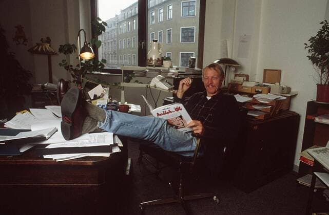 Vito von Eichborn 01 94 am quer ganz sitzend Büro Schreibtisch leger Hemd kariert Jeans lächelnd Mann Unternehmen Verlag Wirtschaft blond Schnäuzer lesend Zeitschrift Telefon