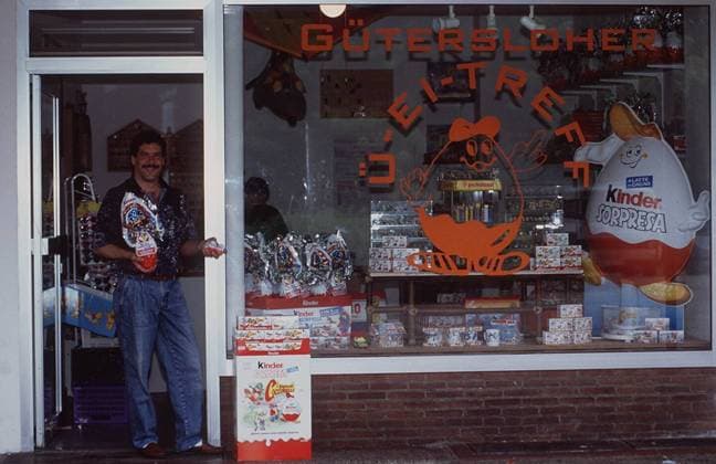 Überraschungs-Ei-Treff mit Gerd Grunert 07 95 her ÜBERRASCHUNGSEI Überraschungseier Mann Sammlung leger Hemd Jeans dunkelhaarig Schnäuzer quer ganz stehend Tür Geschäft draußen Ei haltend lächelnd