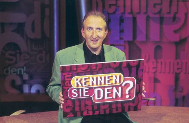 Mike Krüger in Kennen Sie den? (RTL) 06 97 rs Mann TV Schauspiel Show Moderation Film Kino Komik KABARETT dunkelhaarig Halbglatze leger Hemd schwarz Jackett sitzend Studio Dekoration Kulisse Schild Logo haltend halb quer lächelnd