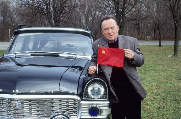 Peter Sodann mit Tschaika in auto motor und sport tv (VOX) 02 00 böw Mann Schauspieler Schauspiel TV Fernsehen Serie dunkelhaarig kurzhaarig Halbglatze leger Jackett grau Pullover Hose schwarz Hemd kariert stehend draußen Wiese Straße Straßenrand angelehnt aufstützend Auto PKW schwarz Tschaika russisches Fabrikat Modell russische Fahne Flagge haltend festhaltend straffziehend am Kühler am Scheinwerfer-Spoiler halb quer nachdenklich lächelnd Fototermin Peter Sodann mit Tschaika am 04.02.2000
