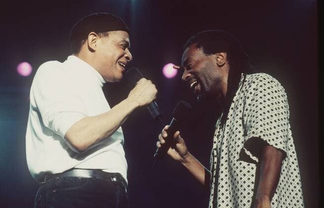 Al Jarreau und Bobby McFerrin (re.) 07 00 thg Mann Musiker Musik Jazzmusiker Jazz Gesang Sänger dunkelhäutig dunkelhaarig leger stehend Saal Halle dunkel Bühne Auftritt halb quer singend sich anschauend Profil Mikrophon haltend Froschperspektive Fototermin Al Jarreau und Bobby McFerrin am 20.07.2000 in der Preussag-Arena in Hannover McFerrin MacFerrin Mc Mac Ferrin