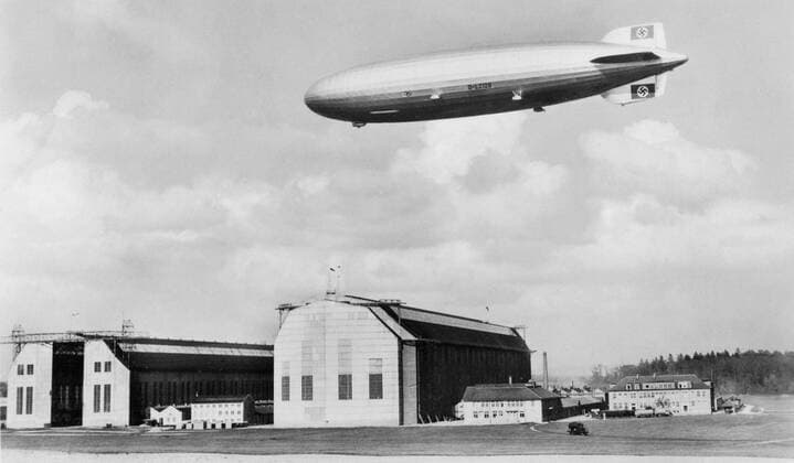 Feature Symbol: Luftschiff Hindenburg 03 10 teuZeppelin LZ 129 Hindenburg ca. 1936 mit Luftschiffhalle in Friedrichshafen am BodenseeDeutschland deutsch historisch historisches Zeppelin Luftschifffahrt Reederei Halle Montagehalle Zeppelinhalle Luftschiffhafen Nazizeit Hakenkreuz Drittes Reich olympische Ringe Olympia Nationalsozialismusgermany historical picture airshipsw quer draußen fahrend schwebend fliegend