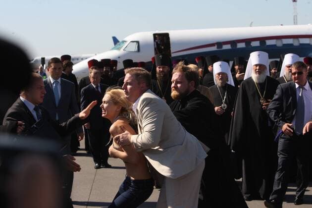 ITAR-TASS: KIEV, UKRAINE. JULY 26, 2012. A security guard detains a topless FEMEN activist as she protests against a visit of Patriarch Kirill of Moscow and All Russia to Ukraine, at Borispol airport.  RE103E10