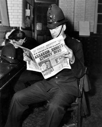 IMAGO History, Ermordung von US-Präsident John F. Kennedy in Dallas, 1963 Policeman reading newspaper - Daily Sketch - with front page headline saying Assassin shoots Kennedy dead - newspaper dated Saturday 23rd November 1963, Zeitung, Titelseite, Schlagzeile, Tageszeitung, Verbrechen, Polizei, Polizist, Polizeimann UnitedArchives00373628 