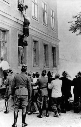 Frau Frieda Schulze makes her dramatic bid to reach freedom - she is pictured hanging from the window of her second storey apartment just inside the West Berlin border . East German police holding one arm try to pull her back . Below a young West German tries to free her to fall into a net held by West German police on the sidewalk , which is in West Berlin . Part of the wall dividing East from West is seen to the right of the house . Her escape was successful . UnitedArchives00935142 