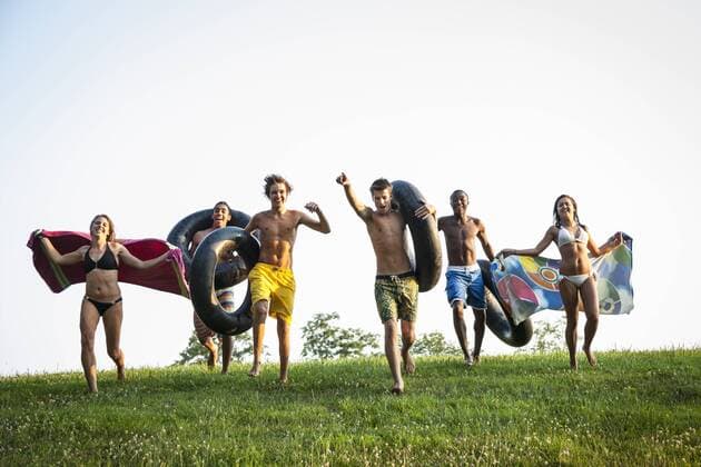 A group of teenagers, boys and girls, running across the grass holding swim towels and inflated floats. 