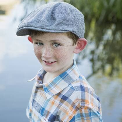 A young boy wearing a checked shirt and cloth cap with a large brim standing on a riverbank. 