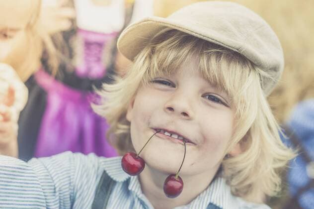IMAGO Creative: Landleben - Essen Portrait of little boy holding two cherries with his mouth model released Symbolfoto  MJF001587