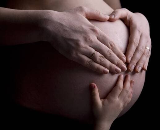 Schwangere in der 29. Woche haelt sich ihren Bauch, formt dabei mit ihren Haenden ein Herz, Kinderhand liegt auch auf dem Bauch 29-weeks pregnant belly with mothers hands forming heart shape and small kids hand BLWS237830