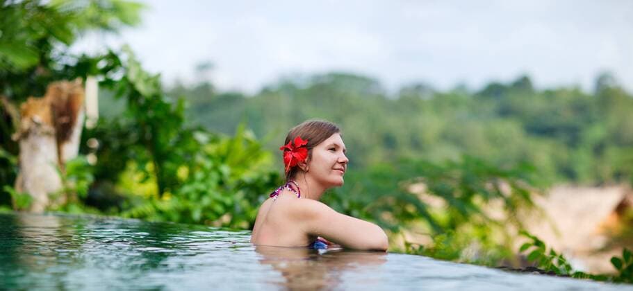 eine junge Frau mit roter Bluete im Haar entspannt sich im Swimmingpool und geniesst die Aussicht a young woman with red flower in her hair relaxing in swimming pool and admiring the view BLWS270739