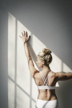 A blonde woman, in a white crop top and leggings, standing in front of a white wall, doing yoga, her arm raised, touching the wall. A blonde woman, in a white crop top and leggings, standing in front of a white wall, doing yoga, her arm raised, touching the wall. 16684