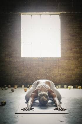 A blonde woman in a white crop top and leggings, bending down on a yoga mat surrounded by candles, doing yoga. A blonde woman in a white crop top and leggings, bending down on a yoga mat surrounded by candles, doing yoga.  16696