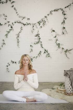 A blonde woman in a white leotard and leggings, sitting on a white mat in a room, doing yoga. A creeper plant on the wall behind her. A blonde woman in a white leotard and leggings, sitting on a white mat in a room, doing yoga. A creeper plant on the wall behind her.  16722
