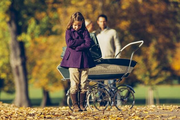 Little girl with crossed arms standing in front of a baby pram in a park with parents in background model released Symbolfoto  CHAF001079