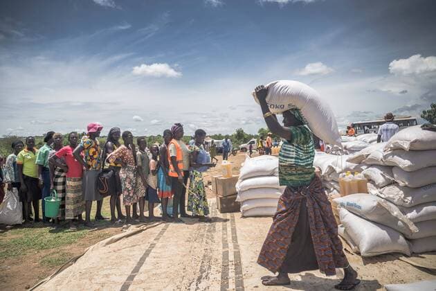 May 7, 2018 - Bidi Bidi, Uganda - A female South Sudanese refugees seen carrying a pack of 50 KG of cassava flour on top of the her head while others line up to get their food supplies..The Bidi Bidi refugee settlement in northern Uganda near the South Sudanese border is currently the largest refugee camp in the world, hosting over 250,000 South Sudanese refugees fleeing the conflict. The world food program is providing monthly basic food supply for the refugees in the settlement. Bidi Bidi Uganda - ZUMAs197 20180507_zab_s197_004 Copyright: xGeovienxSox