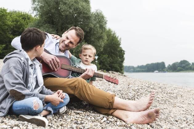 Happy father with two sons sitting at the riverside playing guitar model released Symbolfoto  UUF13955