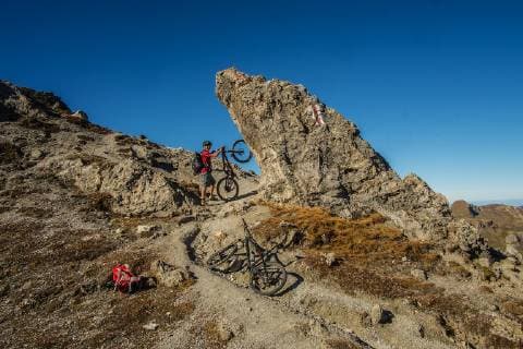 Ein Mountainbikder steht mit seinem Bike an einem markanten Fels am Aufstieg kurz vor der Passhöhe des Wissmilen Pass (2420 m) auf dem Gebiet Flumserberg, Kanton St. Gallen, Schweiz. MTB Tour in den Schweizer Alpen.