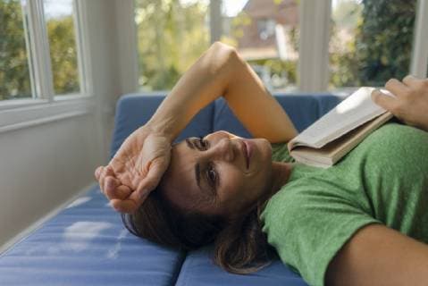 IMAGO Creative: Internationaler Frauenmonat, Gesundheit, Frauen in der Menopause Smiling mature woman lying on couch at home with book model released Symbolfoto property released  KNSF04626