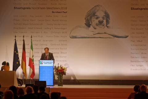 Verleihung des Staatspreises des Landes NRW 2018 an Bernhard Paul. Im Bild der Ministerpräsident des Landes Nordrhein- Westfalen am Rednerpodium auf der Bühne während seiner Ansprache. Im Vordergrund sind Gäste zu sehen. Hinter Laschet prangt an der Rückwand eine Grafik von Bernhard Paul als Clown. Köln Nordrhein-Westfalen Deutschland