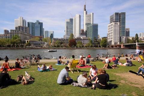 Frühlingstag am Ostermontag in Frankfurt: Sonnenhungrige genießen die warmen Temperaturen und die Sonnenstrahlen bei blauem Himmel auf einer Liegewiese am Mainufer vor den Hochhäusern des Bankenviertel s in der Innenstadt, Hessen, Deutschland