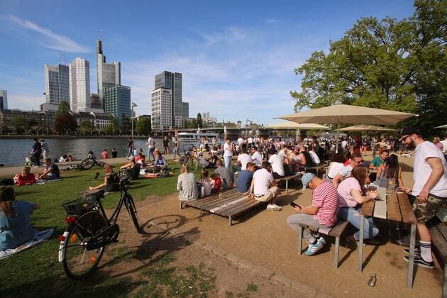 Frühlingstag am Ostermontag in Frankfurt: Sonnenhungrige genießen die warmen Temperaturen und die Sonnenstrahlen bei blauem Himmel auf einer Liegewiese mit Biergarten am Mainufer vor den Hochhäusern des Bankenviertel s in der Innenstadt, Hessen, Deutschland