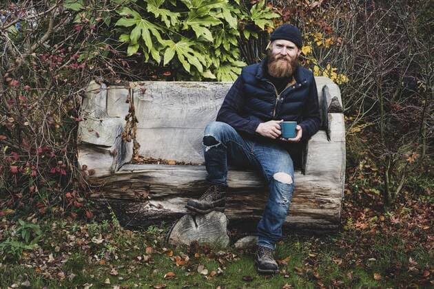IMAGO Creative, Jahreszeit Herbst auf dem Land Bearded man wearing black beanie sitting on wooden bench in garden, holding blue mug, looking at camera. Bearded man wearing black beanie sitting on wooden bench in garden, holding blue mug, looking at camera.  53358