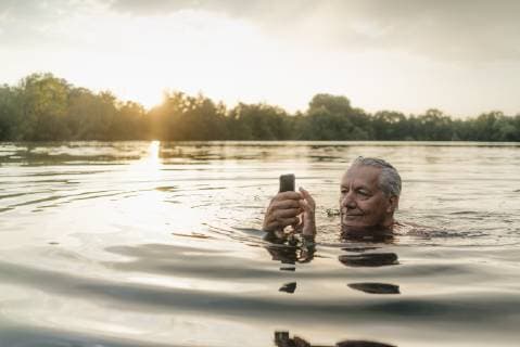 Senior man swimming in a lake at sunset using cell phone model released Symbolfoto  GUSF01823