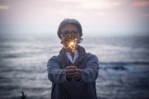 IMAGO Creative: Guten Rutsch Happy senior woman standing in front of the sea by sunset holding sparkler model released Symbolfoto  SIPF01980