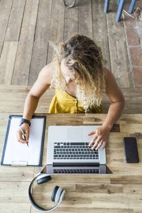 Young woman at wooden desk using laptop and writing on clipboard model released Symbolfoto property released  TCF06171