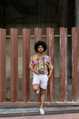 Man with a camera wearing colorful shirt leaning against a fence model released Symbolfoto  AFVF03873