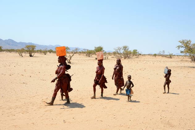 Himba women and children returning from the waterpoint, carrying plastic cans full of water, during dry season, Marienfluss Valley, Kaokoland Desert, Namibia. October 2015  1531101 EricxBaccega