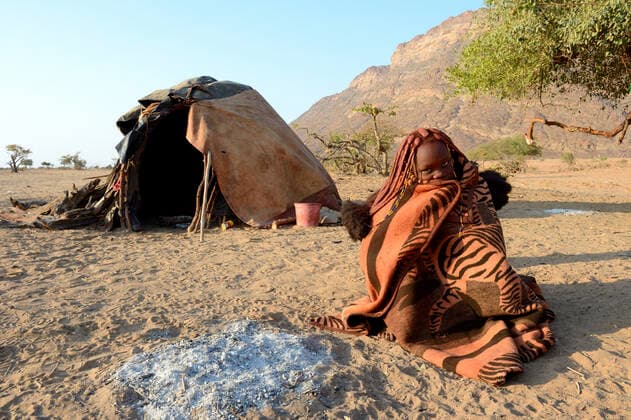 IMAGO Cultures of the world, Himba Volk in Namibia Himba woman wrapped up in a blanket next to her open fire with her mud hut in the background. Marienfluss Valley. Kaokoland, Namibia October 2015  1531138 EricxBaccega