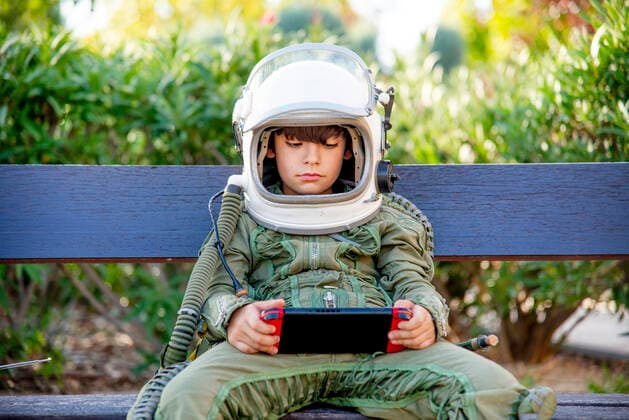 IMAGO Creative, Lifestyle, Gaming Creative Highlights Symbolfotos Boy wearing a space suit and sitting on a bench, playing alone with video games model released Symbolfoto  CJMF00124