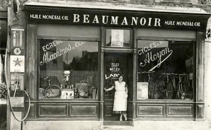 CYCLISME annees 10 Devanture du magasin de cycles et de motos Beaumanoir avec pompe a essence, patronne devant la porte. Photographie anonyme pour une carte postale vers 1920 1930. Credit : Collection IM KHARBINE-TAPABOR. *** CYCLING in the 10s Front of the Beaumanoir bike and motorcycle shop with gas pump, boss in front of the door Anonymous photograph for a postcard around 1920 1930 Credit Collection IM KHARBINE TAPABOR  KH177087