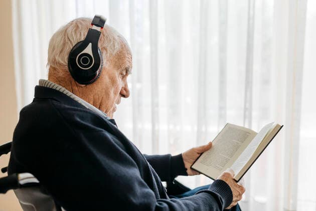 Senior man sitting in wheelchair reading a book while listening music with headphones model released Symbolfoto property released  JRFF03946