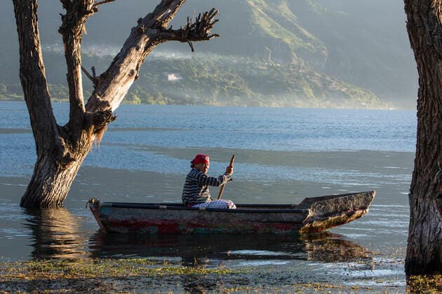 Mayan man in the traditional dress of San Pedro la Laguna paddles a cayuco in early morning light on Lake Atitlan, Guatemala. Side view, opposite lake shore and town of San Pablo la Laguna visible in distance. Cayuco is between two standing dead trees in the water. Lake level has risen more than 30 since 2009, flooding shores and houses and killing these San Pedro la Laguna Solola Guatemala Copyright: xJonxG.xFullerx JGF-2735